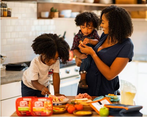 A mother smiling and holding a young child while another child prepares food on the kitchen counter, surrounded by fresh ingredients
