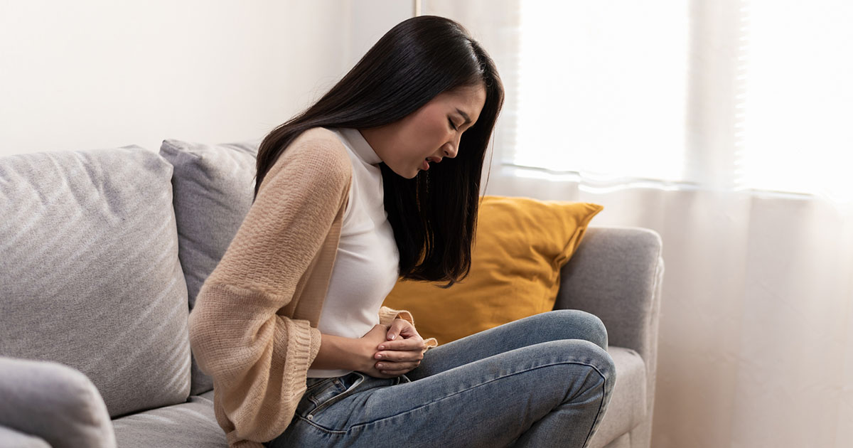 A young Asian woman sitting on a sofa, clutching her stomach in pain, likely experiencing discomfort such as stomach ache, cramps, or menstrual pain.